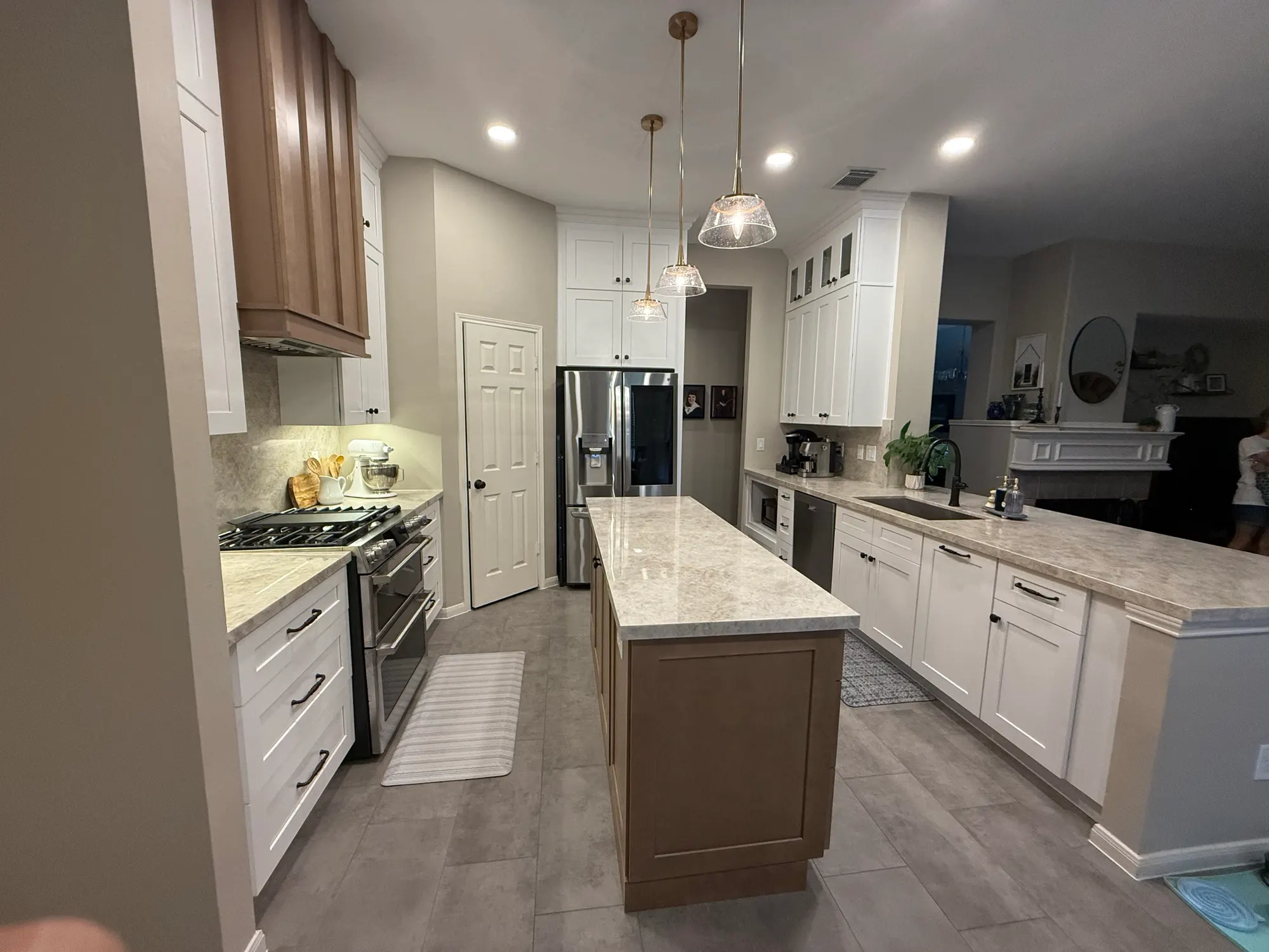 After: the same kitchen with white shaker cabinets to the ceiling, a stained wood range hood and island, quartzite counters and new gray tile floors